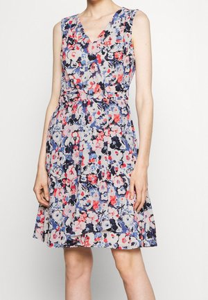 Mouwloze jurk tot op de knie met een bloemenpatroon in roze, blauw, zwart en wit en een wikkelstijl bovenlichaam, gedragen door een persoon die staat tegen een effen witte achtergrond.