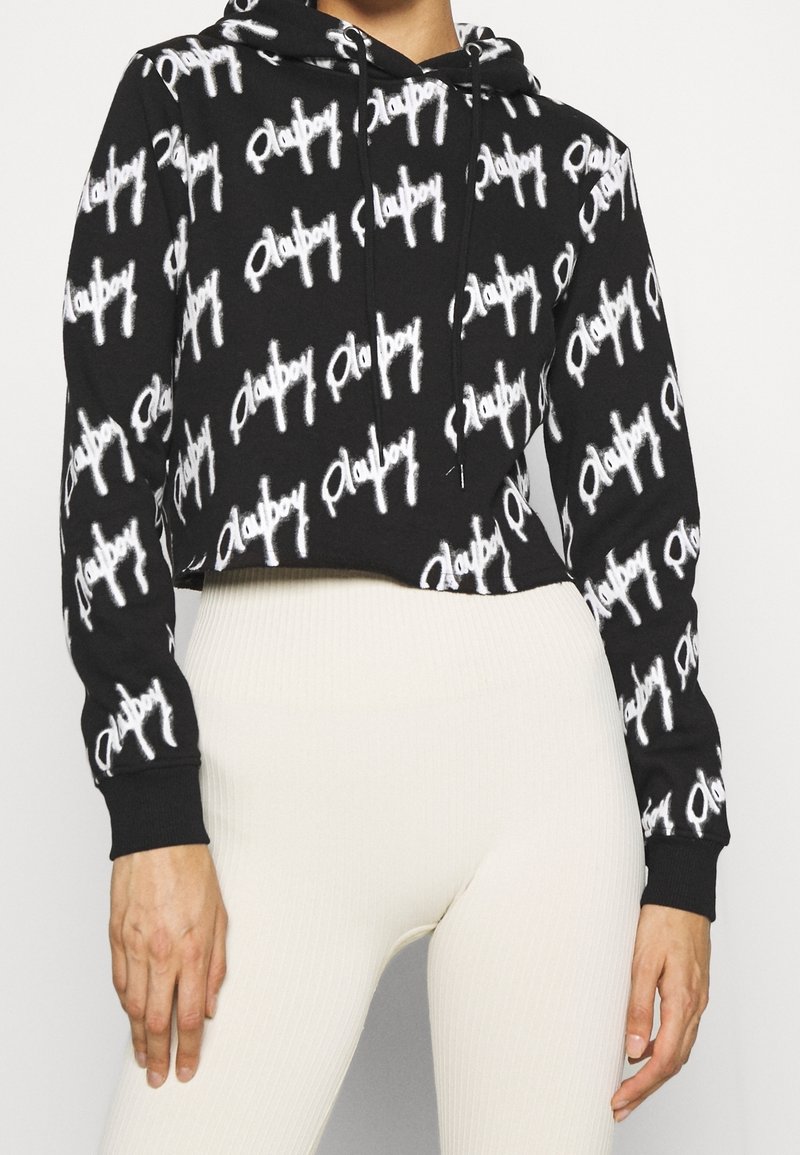 Hoodie noir court avec le script "Playboy" blanc répétitif, associé à un legging côtelé taille haute de couleur blanc cassé, porté par une personne debout.