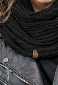 Bufanda infinita negra de punto con diseño texturizado y acanalado, que presenta una pequeña etiqueta de cuero marrón. Llevada sobre una chaqueta de cuero negra.