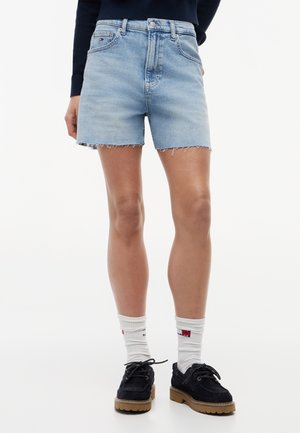 Γυναίκα που φοράει κοντά παντελόνια από ανοιχτό μπλε τζιν, λευκές κάλτσες με λογότυπο και μαύρα δερμάτινα παπούτσια με κορδόνια, σε λευκό φόντο.