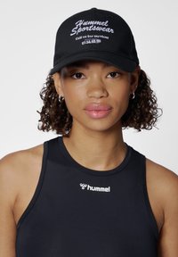 Schwarze Kappe mit "Hummel Sportswear" in weißer Schrift gestickt. Strapazierfähiger Stil, getragen über einem schwarzen ärmellosen Oberteil mit Logodetails.
