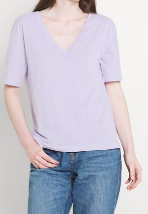 Vrouw draagt een lichtpaars V-hals topje met korte mouwen en een blauwe spijkerbroek, met de ene hand op haar heup en de andere ontspannen langs haar zijde.