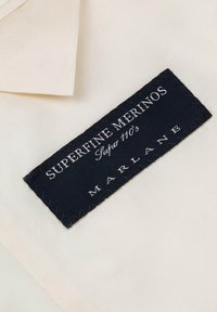 Biała tkanina z ciemnoniebieską metką wszytą w nią. Metka ma napis "Superfine Merinos" oraz "Marlane" w białym kolorze.