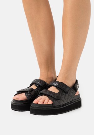 Black quilted platform sandals with dual hook-and-loop straps, flat rubber sole, and open toe design. Smooth texture and minimal hardware detail.
