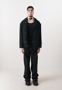 Veste noire avec poches, portée sur une chemise noire, assortie avec un pantalon noir usé et des bottes noires. Design simple et utilitaire.