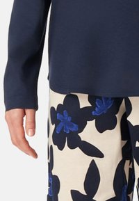 Person trägt ein marineblaues langärmeliges Oberteil und cremefarbene Hosen mit großem marineblauem und blauem Blumenmuster, Hand entspannt an der Seite.