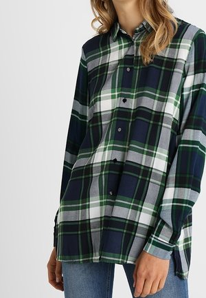 Kobieta ubrana w koszulę na guziki z długim rękawem w zieloną, granatową, białą i szarą kratę, zestawioną z niebieskimi dżinsami.