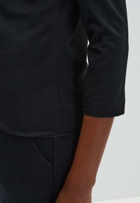 Haut noir à manches longues avec une texture lisse, présentant un ourlet arrondi et des coutures visibles près de la couture latérale, associé à un pantalon sombre à texture.