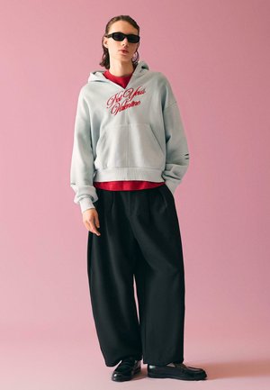 Persona con occhiali da sole neri, felpa con cappuccio grigio chiaro con scritte rosse, pantaloni neri larghi, scarpe nere, in piedi davanti a uno sfondo rosa.