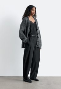 Graue Mohair-Strickjacke, schwarzes Spitzen-Camisole, hochtaillierte schwarze Hosen mit weitem Bein und flache schwarze Schuhe, vor neutralem Hintergrund.