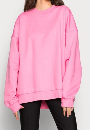 Femme portant un sweat-shirt oversize rose vif à manches longues et un pantalon noir, debout devant un fond clair uni.