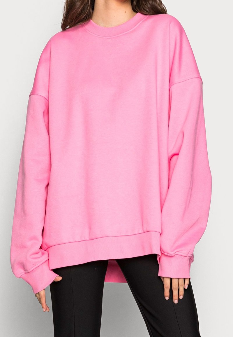 Femme portant un sweat-shirt oversize rose vif à manches longues et un pantalon noir, debout devant un fond clair uni.