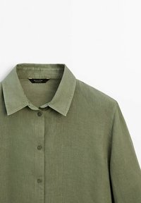 Chemise en lin vert olive à boutons, avec un col classique, des manches longues et des coutures visibles sur un fond blanc.