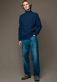 Marineblauer Strickpullover mit Rollkragen und strukturiertem Muster, kombiniert mit hellblauen, lässig geschnittenen Jeans und grauen Schuhen vor einem neutralen Hintergrund.
