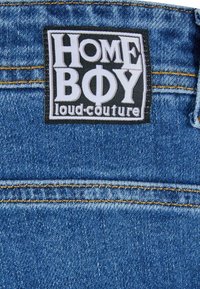 Plava traper tkanina s crnom i bijelom oznakom na kojoj piše "HOME BOY loud.couture." Ima žute šavove i teksturiranu površinu.