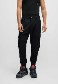 Pantalones cargo negros con un diseño ajustado, que cuentan con bolsillos laterales y un cierre de botón. Combinados con zapatillas negras con detalles rojos.