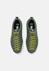 Zielone dzianinowe sneakersy z żółtymi sznurówkami, charakteryzujące się oddychającą cholewką i białą gumową podeszwą. Logo umieszczone na języku.