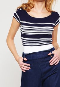 Marinblå randig stickad topp med vita accenter och korta ärmar. Har knapppdetaljer på axlarna. I kombination med högmidjade marinblå byxor.