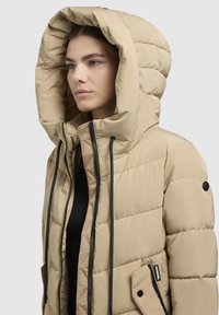 Doudoune beige avec une grande capuche, dotée de zips noirs et d'une poche frontale. Le tissu a une texture matelassée et un design épuré.