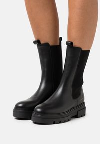 Par de botas negras de cuero hasta el tobillo con suelas de goma gruesa, utilizadas sobre piernas desnudas, que cuentan con tiradores y paneles elásticos en los laterales.