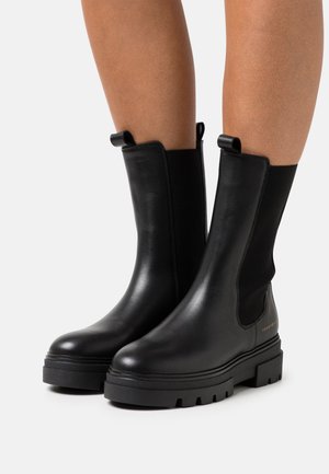 Une paire de bottines en cuir noir avec des semelles en caoutchouc épaisses, portées sur des jambes nues, avec des languettes de tirage et des panneaux élastiques sur les côtés.
