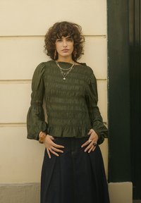 Frau mit lockigem Haar, die eine grüne, langärmlige, geraffte Bluse, mehrere Halsketten und einen dunklen Rock trägt, während sie vor einer beigen Wand steht.