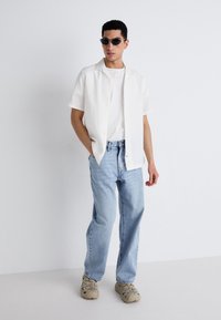 Chemise blanche à manches courtes par-dessus un t-shirt blanc uni, jeans bleu clair et baskets beiges. Modèle portant des lunettes de soleil noires, se tenant de manière décontractée.