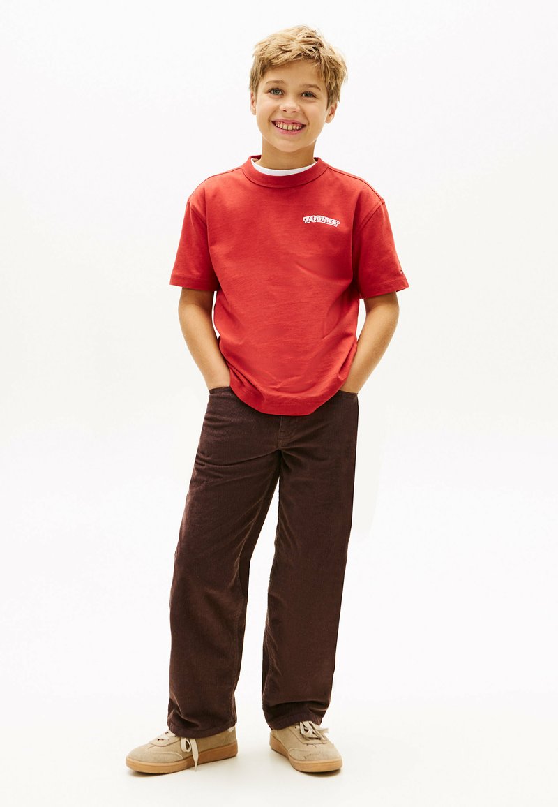 Rød bomulds T-shirt med rund hals og hvidt logo, parret med mørkebrune kordfløjlsbukser. Lyse beige sneakers med snørebånd.