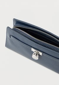 Denim modra usnjena denarnica z zadrgo, teksturirano površino in srebrno kovinsko opremo s krožnim logotipom ter žepom spredaj.