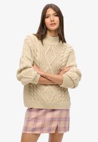 Beige cable-knit sweater with a high neck and thick ribbed hem, worn over a pink plaid mini skirt featuring a front slit.