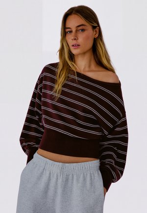 Ung kvinde med langt blondt hår iført en brunt stribet off-shoulder sweater og lysegrå joggingbukser mod en ensfarvet baggrund.