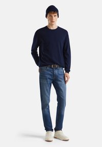 Marineblauer Pullover, Jeans aus Denim, weiße Sneakers und eine marineblaue Strickmütze. Der Pullover hat eine strukturierte Oberfläche, die Jeans zeigen einen leichten Farbverlauf und die Schuhe haben eine glatte Oberfläche.