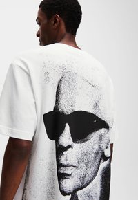 Biely bavlnený tričko s veľkým čiernym grafickým znázornením tváre v slnečných okuliaroch na zadnej strane, so voľným strihom a okrúhlym výstrihom.