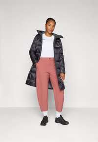 Žena stoji samouvjereno u crnoj puffer jakni, bijeloj majici i ružičastim hlačama, noseći crne tenisice i bijele čarape, ispred sive pozadine.