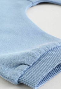 Svetlomodrá pletená látka s rebrovaným manžetovým detailom, zobrazujúca textúru a šitie na hladkom bielom povrchu.