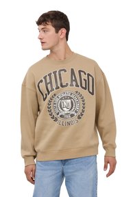 Felpa beige con la scritta "CHICAGO" e grafica del logo di colore marrone scuro. Caratterizzata da collo rotondo a coste, maniche lunghe e una vestibilità comoda.