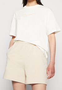 Ljusbeige bomullströja med en diskret orange Nike-logotyp, i kombination med beige shorts som har en liten vit Nike-logotyp på sidan.