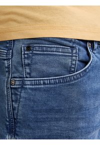 I jeans in denim blu presentano cuciture sui taschini frontali, un leggero effetto di lavaggio e un evidente bottone nero accanto al taschino.