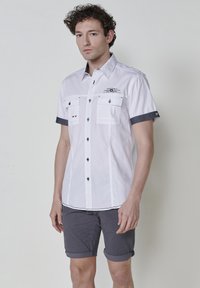 Camisa blanca de manga corta con detalles en azul oscuro, dos bolsillos en el pecho y cierres de botón negros. Llevada con pantalones cortos grises enrollados.