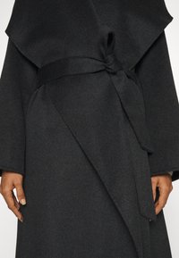 Schwarzer Wollumhang mit weiten Ärmeln, ausgestattet mit einem gebundenen Gürtel in der Taille und einer diagonalen Überlappung an der Front. Weiche Textur, minimalistisches Design.