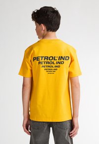 Camiseta de algodón amarilla con mangas cortas, que presenta un gran texto en negro en la parte posterior, que dice "PETROLIND" varias veces.