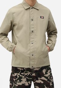 Béžová lehká bunda s předním zapínáním na knoflíky, elastickými manžetami a bočními kapsami. Na hrudi se nachází malá nášivka s logem. Kombinována s maskáčovými kalhotami.