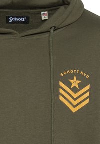 Bluza z kapturem w oliwkowej zieleni o fakturowanej tkaninie. Na piersi znajduje się żółta gwiazda i wzór w kształcie szewrona, kaptur ze sznurkami oraz metka z logo.