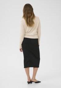 Una blusa de manga larga color beige claro combinada con una falda negra hasta la rodilla. La falda presenta un diseño elegante con dos bolsillos traseros. Mules de tacón negro.