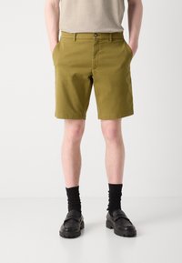 Olivenfargede shorts med klassisk passform, laget av bomull, med beltehemper, sidelommer og knappelukking. Brukt med svarte sokker og sko.