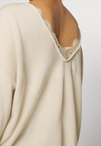 Lichtbeige trui met een V-hals en kantafwerking aan de achterkant. Gebreide stof, zachte textuur, met rafelige randen langs de halslijn.