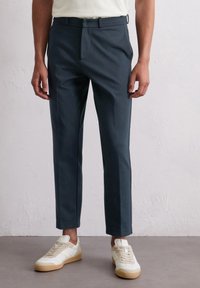 Schmale, navyfarbene Hosen aus glattem Stoff, mit flacher Vorderseite und Gürtelschlaufen, kombiniert mit weißen und hellbraunen Sneakers.