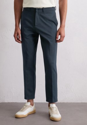 Navy slim-fit spodnie wykonane z gładkiego materiału, z płaskim przodem i pętelkami na pasek, połączone z białymi i beżowymi sneakersami.