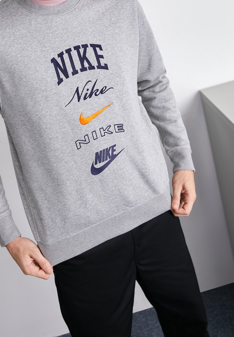 Personne portant un sweat-shirt gris avec plusieurs logos Nike de styles et couleurs différents à l'avant, tenant l'ourlet du sweat-shirt.