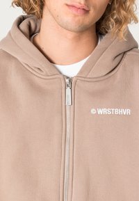 Sudadera con capucha marrón con cremallera, hecha de tela suave con un bolsillo canguro, que presenta un texto de logo blanco en el pecho y forro texturizado en la capucha.